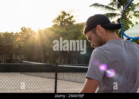 Il giocatore di tennis maschile si trova sul campo da tennis all'aperto con una racchetta sulla spalla e guarda verso il basso. Foto Stock