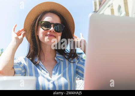 Giovane donna in cappello di paglia e occhiali da sole che posano davanti al suo computer portatile mentre si siede in un bar all'aperto in estate. Foto Stock