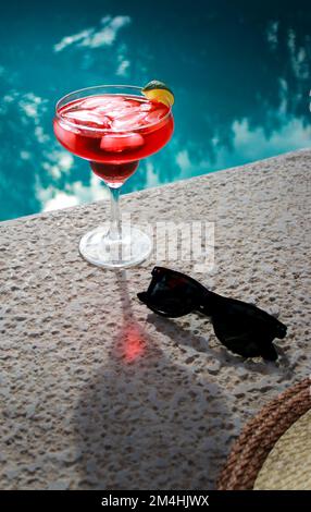 Primo piano di cocktail rosso freddo in piscina con occhiali da sole neri e cappello di paglia Foto Stock