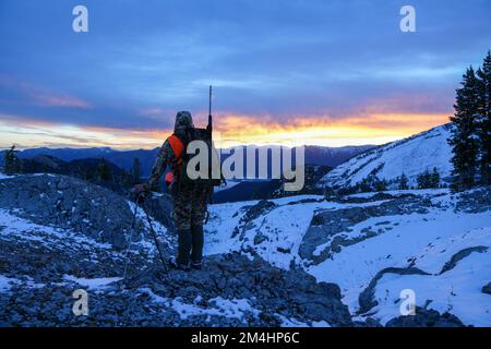 Cacciatore con fucile vestito in camo si erge sul bordo di una cresta innevata di montagna alla ricerca di cervi all'alba nel Wyoming Foto Stock