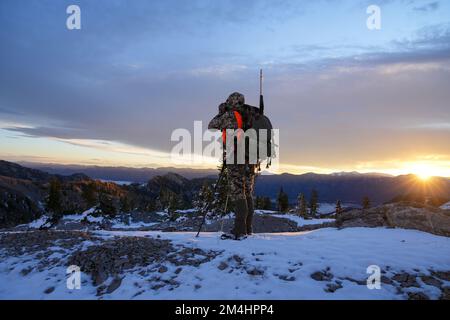 Cacciatore con fucile vestito in camo si erge sul bordo di una cresta innevata di montagna alla ricerca di cervi all'alba nel Wyoming Foto Stock