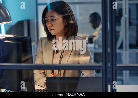 Giovane imprenditrice asiatica in tuta che si concentra sul suo lavoro sul computer nel suo posto di lavoro in ufficio Foto Stock