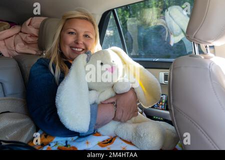 Wyoming, USA - 20 luglio 2022: Donna bionda felice si siede sul retro di un'auto, tenendo e abbracciando un coniglietto di animali farciti Foto Stock
