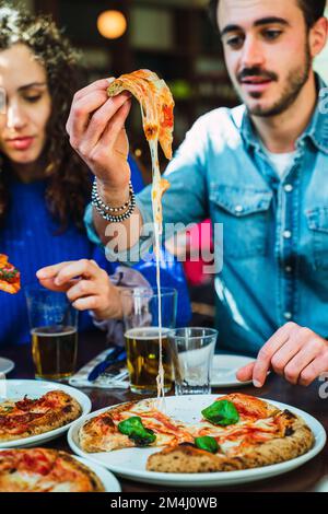 Ragazzo prendere una fetta di pizza fuori dal piatto. Il formaggio stretching e apparente appetitoso. Amici che gustano pizza al formaggio con crosta croccante. Bicchiere di birra Foto Stock