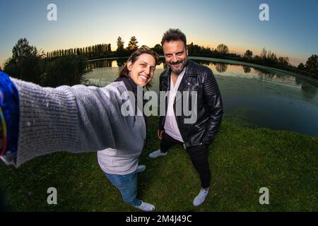 Coppia di amici che prendono un selfie al tramonto vicino ad un lago mentre sorridono felicemente. Concetto di gioia, felicità e spensieratezza. Effetto fotocamera fisheye Foto Stock