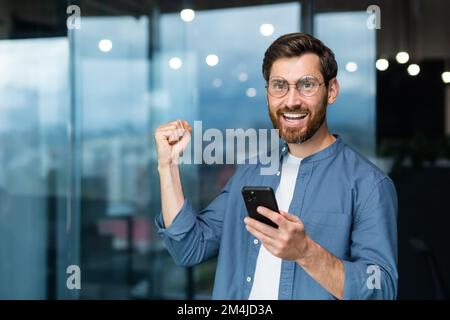 Ritratto di un uomo d'affari moderno nel mezzo dell'ufficio, l'uomo sta guardando la fotocamera con uno smartphone e celebrando la vittoria, l'uomo sta tenendo il telefono nelle sue mani e alzando la mano in un gesto di trionfo. Foto Stock