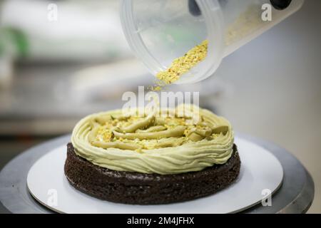 designer di pasticceria che usa crema di pistacchio e spruzzare su strati di torta di cioccolato smerigliato scuro al laboratorio di cucina Foto Stock