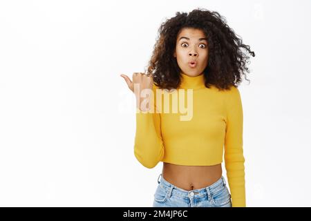 Sorpresa nera ragazza giovane donna che guarda stupito e puntando a sinistra al testo promo, spazio vuoto copia, dire wow, in piedi su sfondo bianco Foto Stock