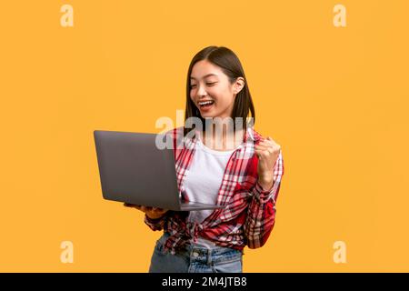 Ritratto di felice giovane donna asiatica che tiene il portatile e gesturing sì, scuotendo pugno clenched, leggendo grandi notizie Foto Stock