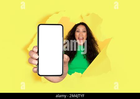 Giovane donna overjoyed che mostra lo smartphone moderno con schermo vuoto attraverso il foro lacerato nella parete di carta gialla, mockup Foto Stock