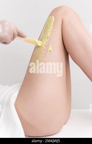 Mano nel guanto applicando cera calda verde sulla gamba della donna sottile utilizzando la spatola. Procedura di depilazione con cera calda in salone di bellezza Foto Stock