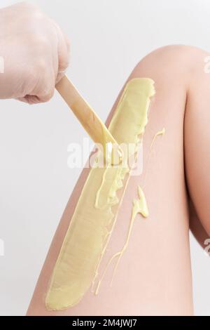 Mano di primo piano nel guanto che applica la cera calda verde sulla gamba della donna usando la spatola. Procedura di depilazione professionale con cera calda in salone di bellezza Foto Stock