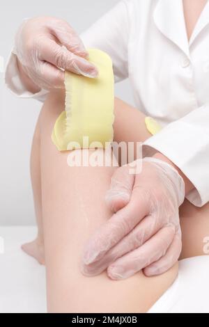 Procedura di depilazione - cosmetologa mani in guanti rimuovendo i capelli sulle gambe delle donne. Processo di ceratura con cera calda verde in salone di bellezza professionale Foto Stock