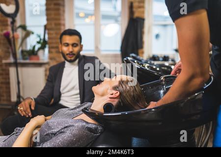 barbiere lavare i capelli della donna al salone e suo marito ora di attesa sul divano. Foto di alta qualità Foto Stock