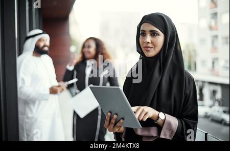 Potrebbe funzionare... una giovane e attraente donna d'affari vestita con abiti tradizionali islamici usando un tablet sul balcone del suo ufficio. Foto Stock