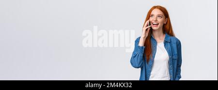 Amichevole spensierata bella donna redhead parlare al telefono, avendo interessante discussione con l'amico sullo smartphone, chiamando mamma che vive all'estero, in piedi Foto Stock