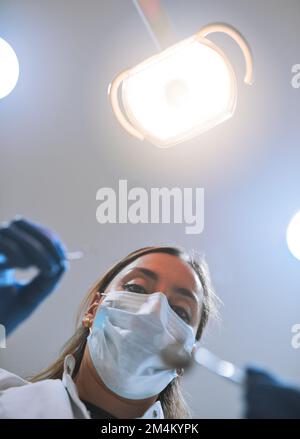 Aperto completamente. Ripresa pov ad angolo basso di una giovane dentista focalizzata che indossa una maschera chirurgica mentre tenta di lavorare sui denti di un paziente. Foto Stock