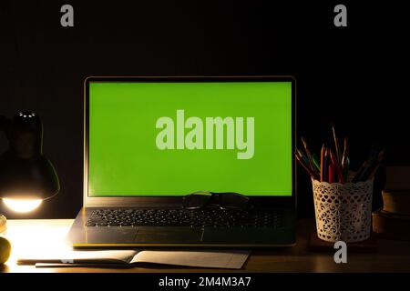 computer portatile con schermo verde sul tavolo con libri vicino alla lampada luminosa inclusa nella camera da letto buia a casa Foto Stock