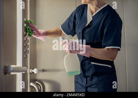 Primo piano della cameriera che indossa uniforme e guanti pulizia ascensore con detergente e straccio Foto Stock