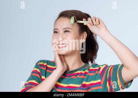 Giovane donna allegra che guarda lo specchio e sorridente soddisfatto mentre massaggia la guancia con un rullo per il viso in giada Foto Stock