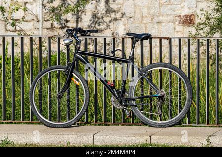 Bicicletta nera parcheggiata e incatenata a una recinzione in un parco con vegetazione verde su un muro di pietra sullo sfondo Foto Stock