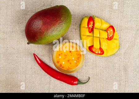 Vista dall'alto di mango intero, peperoncino intero, pepe caldo tagliato a fette, metà di mango tagliata e chutney di mango piccante in un recipiente di vetro sul burlap. Foto Stock
