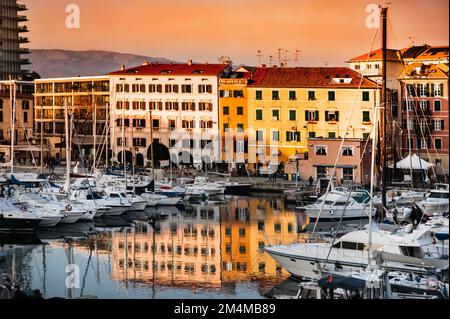 Italia, Savona. Vista, panorama del porto di Savona al tramonto. Barche, yacht e le vecchie case tipiche si riflettono nell'acqua. Foto Stock