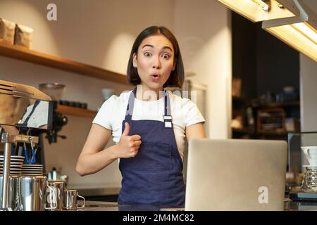 Sorpresa barista ragazza, ragazza caffè mostra pollici in su, consiglia smth, sembra stupito e impressionato, si alza al banco con il laptop Foto Stock