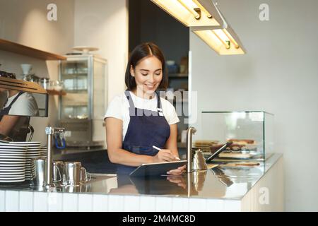 Ritratto di bella ragazza asiatica sorridente, barista in caffetteria che lavora dietro il bancone, utilizzando il tablet come terminale POS, l'elaborazione dell'ordine Foto Stock