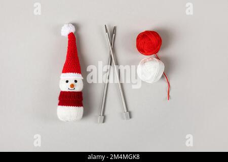 Pupazzo di neve lavorato a maglia in un cappello rosso di Babbo Natale e maglione con due palle di filo e aghi da maglieria su sfondo grigio. Decorazioni di Natale a maglia Foto Stock