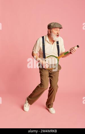 Ritratto di anziano, vecchio in abiti eleganti che giocano con racchetta come chitarra su sfondo rosa. Tempo libero Foto Stock