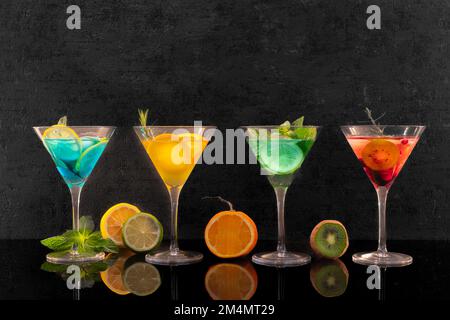 Quattro diversi bicchieri da cocktail con cocktail colorati su sfondo nero e frutta al banco. Foto Stock