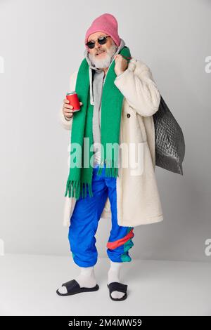 Ritratto di elegante anziano, vecchio in pelliccia cappotto, cappello rosa lavorato a maglia e sciarpa verde su sfondo grigio Foto Stock