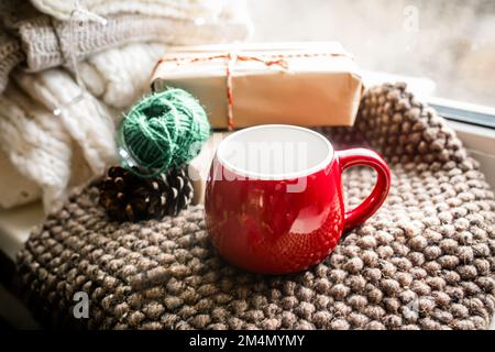 Una tazza rossa vuota di Natale su una coperta accogliente sul davanzale vicino alla finestra. Confezione regalo con filo naturale e carta riciclata. Foto Stock