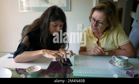 Figlia adulta che spiega le informazioni alla madre anziana utilizzando un dispositivo cellulare. Persona che mostra lo smartphone a una donna anziana Foto Stock