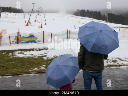 22 dicembre 2022, Sassonia, Oberwiesenthal: Due vacanzieri con ombrelloni si trovano sotto la pioggia sulla pista sciistica di Fichtelberg. Solo pochi giorni dopo l'inizio, la stagione sciistica in Sassonia è minacciata di una pausa più lunga. Sul Fichtelberg, un solo ascensore era aperto lo stesso giorno. Se continua a piovere così, le operazioni saranno sospese da venerdì (23,12.). Precipitazioni e vento stanno mettendo una tensione sulla copertura di neve sulle piste. Secondo il servizio meteorologico tedesco, rimarrà piovoso nei giorni di Natale con temperature fino a dieci gradi, e anche in montagna è ex Foto Stock