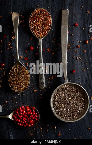 Vecchi cucchiai di metallo con diversi tipi di semi e spezie su sfondo nero. Giacitura piatta. Vista dall'alto. Concetto di cibo. Fotografia di cibo di umore scuro. Foto Stock