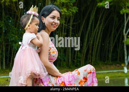 madre latina seduta, con il suo bambino brunetta in piedi al fianco, curiosamente osservando la natura sorridendo, in una foresta di bambù. Foto Stock