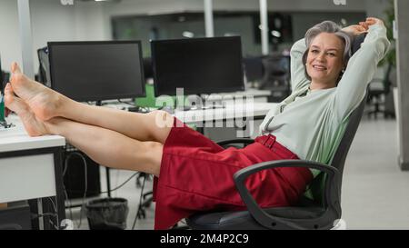 Donna caucasica matura tolse le scarpe e mise i piedi sul desktop in ufficio. Foto Stock