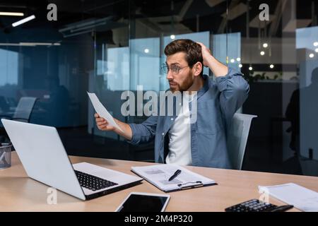 Problemi sul lavoro. Preoccupato e nervoso giovane uomo d'affari maschio, freelancer scioccato, tenendo documenti, fatture nelle sue mani. tenendo la testa con la mano. Seduto in ufficio a un tavolo con un computer portatile. Foto Stock