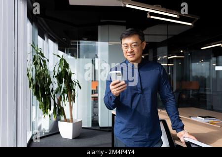 Uomo d'affari asiatico all'interno dell'ufficio in una camicia casual e occhiali sta utilizzando il telefono, il capo sta tenendo uno smartphone nelle sue mani leggere le notizie e digitare un messaggio sorridente. Foto Stock