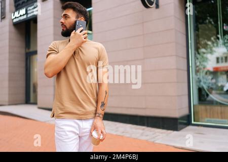 Elegante e bello giovane uomo con mano tatuata che cammina per la strada cittadina, che parla sullo smartphone e che beve un caffè da asporto guardando indietro. Foto Stock