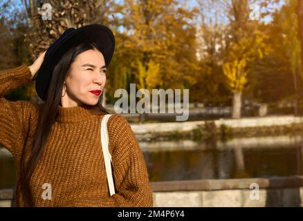Primo piano ritratto di ragazza in un cappello nero e marrone occhio cappotto strinca la macchina fotografica e sorride mentre tiene bianco bag.Sun splendente nell'angolo Foto Stock