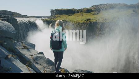 Donna in piedi sul bordo della scogliera guardando la potente cascata di Defoss in Islanda Foto Stock