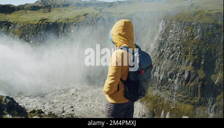 Uomo escursionista in giacca gialla e zaino in piedi sul bordo della scogliera che guarda giù per il canyon con il fiume di montagna dalla cascata di Detifoss in Islanda Foto Stock