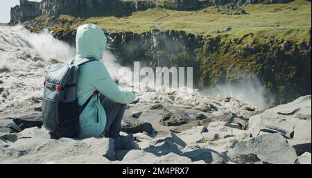 Donna seduta sul bordo della scogliera godendo della cascata di Defoss in Islanda Foto Stock