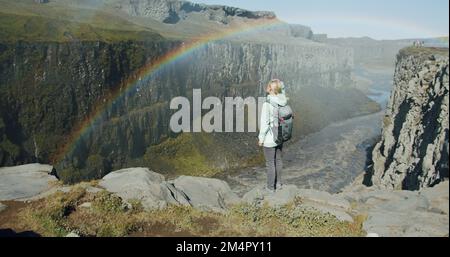 Donna in piedi sul bordo della scogliera che guarda giù per il canyon con il fiume vicino alla cascata di Detifoss in Islanda Foto Stock