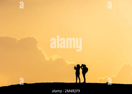 silhouette di una coppia, donna e uomo, in piedi su una collina scattando foto. Luminoso cielo arancione tramonto con alte nuvole edificio sullo sfondo. Foto Stock