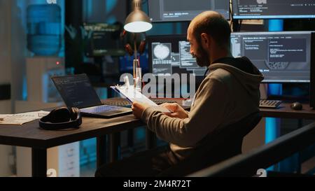 Sviluppatore di software che analizza i dati Web su fogli e schermate degli appunti, confrontando le informazioni sui file. Giovane uomo che usa il linguaggio di programmazione e codifica la nuova interfaccia utente di notte. Foto Stock