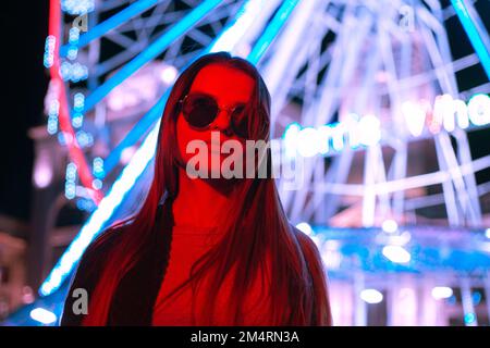 Ritratto una giovane donna urbana piuttosto turistica in occhiali da sole in piedi di fronte ad una ruota panoramica su sfondo rosso al neon di colore chiaro Foto Stock